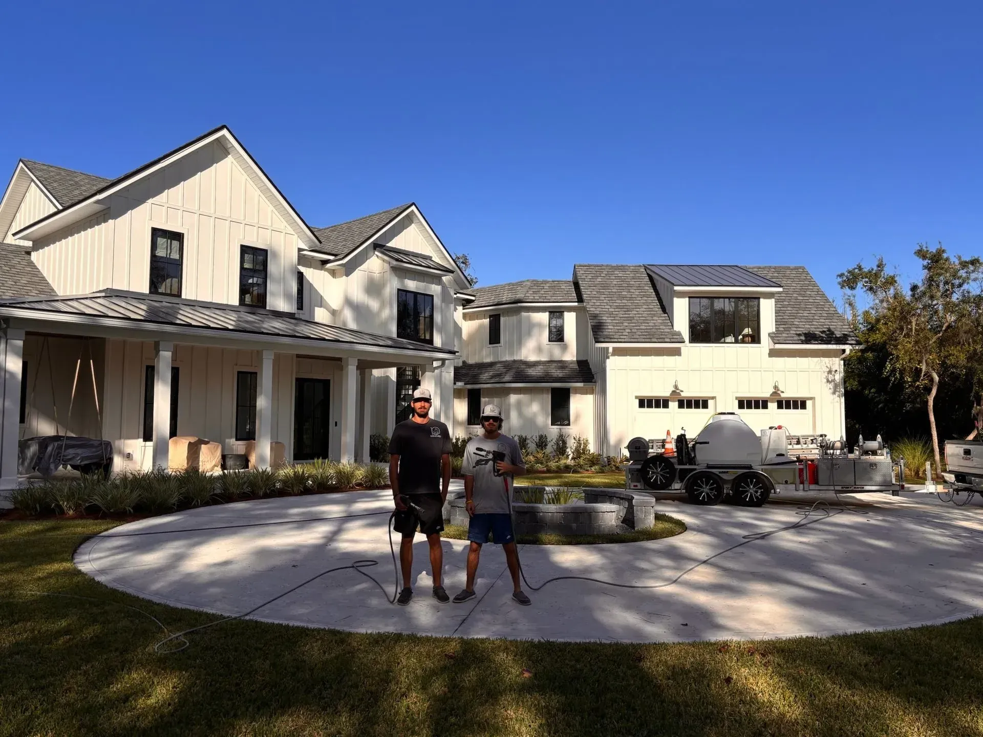 Two men stand in front of a large white house with a circular driveway. Blue sky.