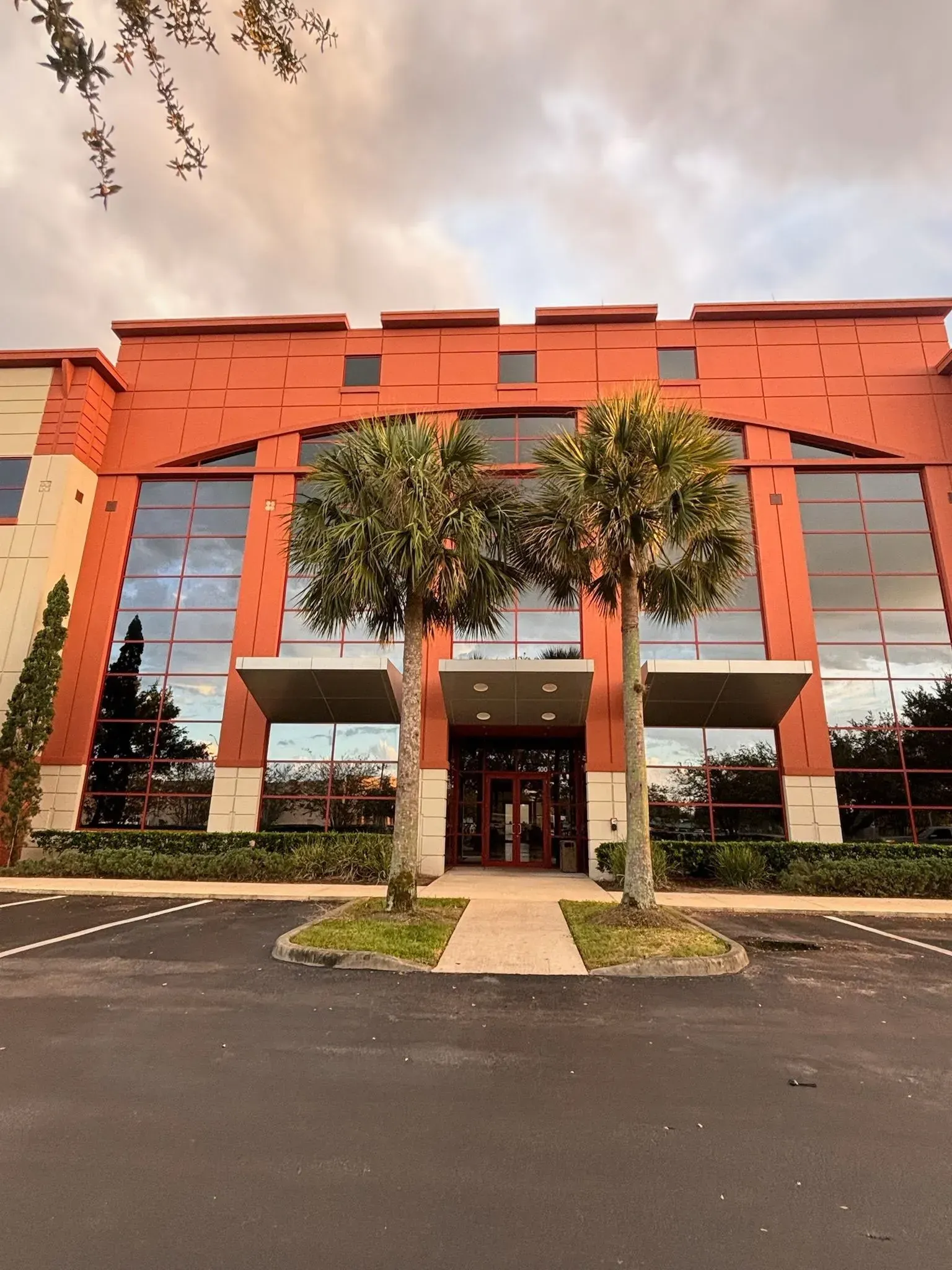 Modern building with red brick facade, large windows, and palm trees at entrance.