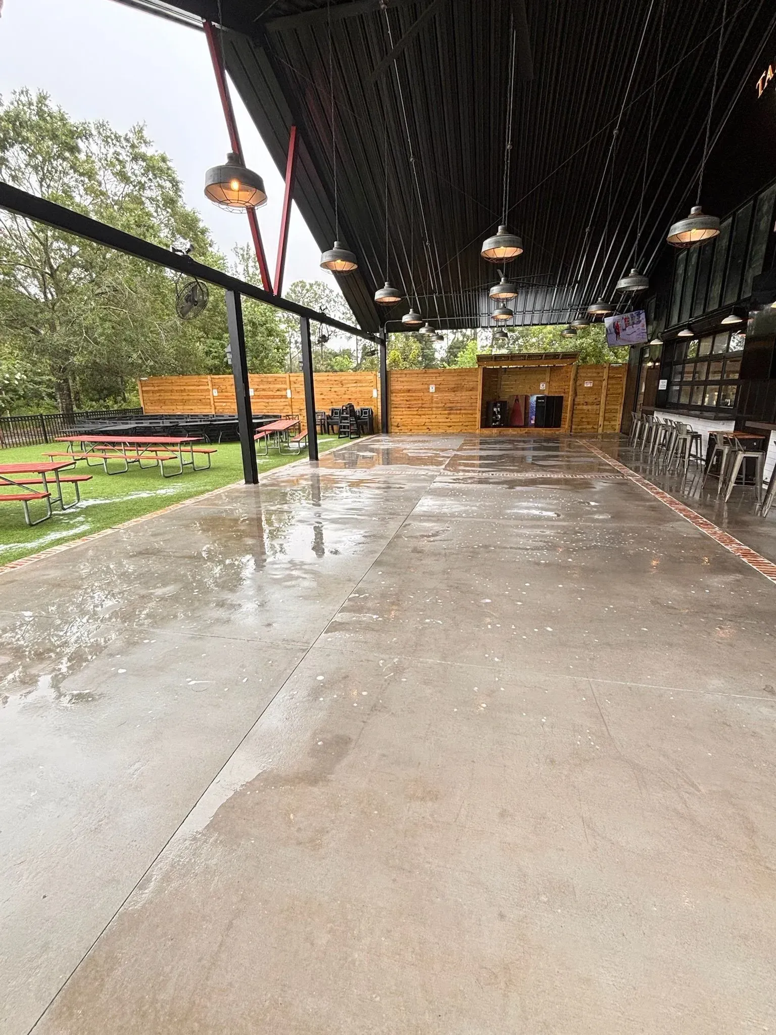 Outdoor patio with wet concrete floor. Tables, string lights, and a bar area are visible.