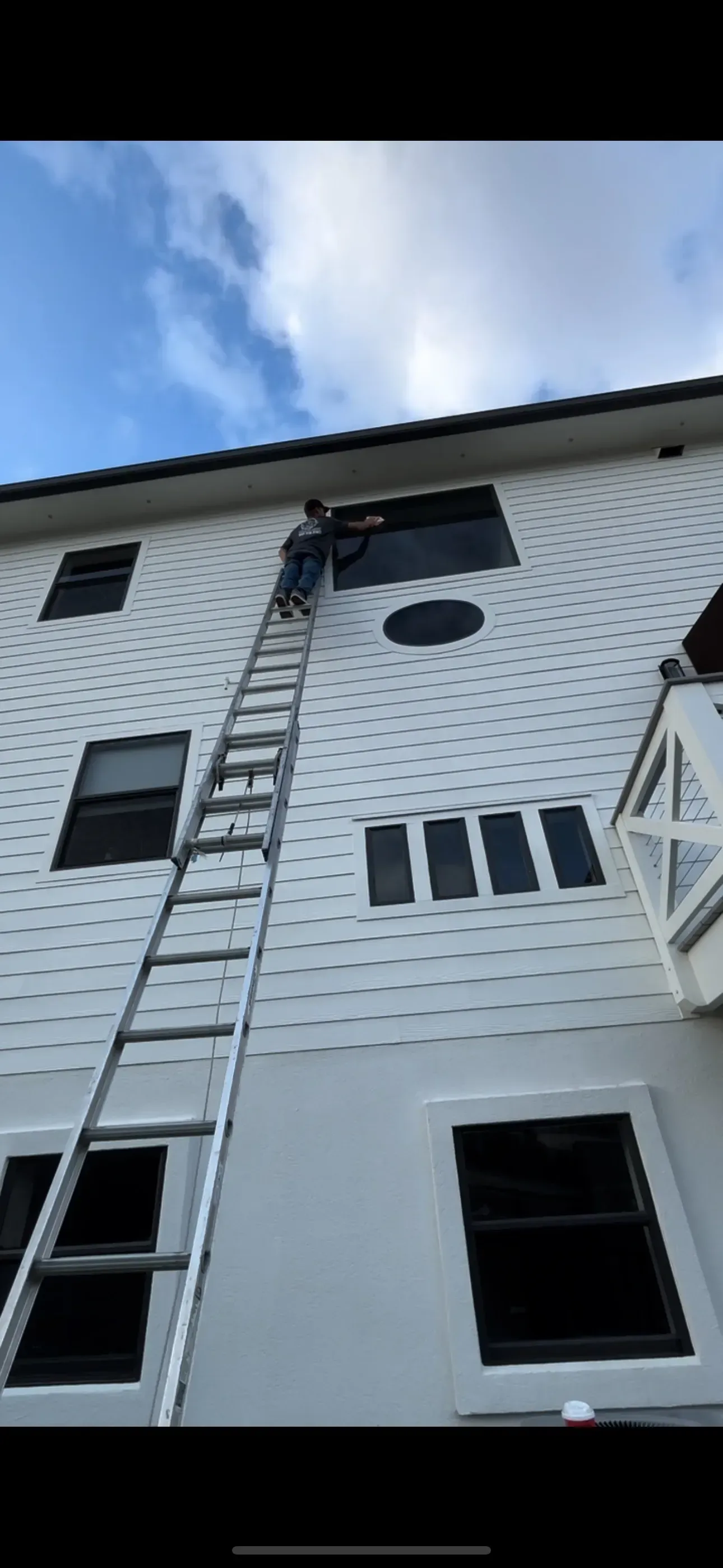 A person climbing a ladder to access a window on the side of a white building with patterned siding.