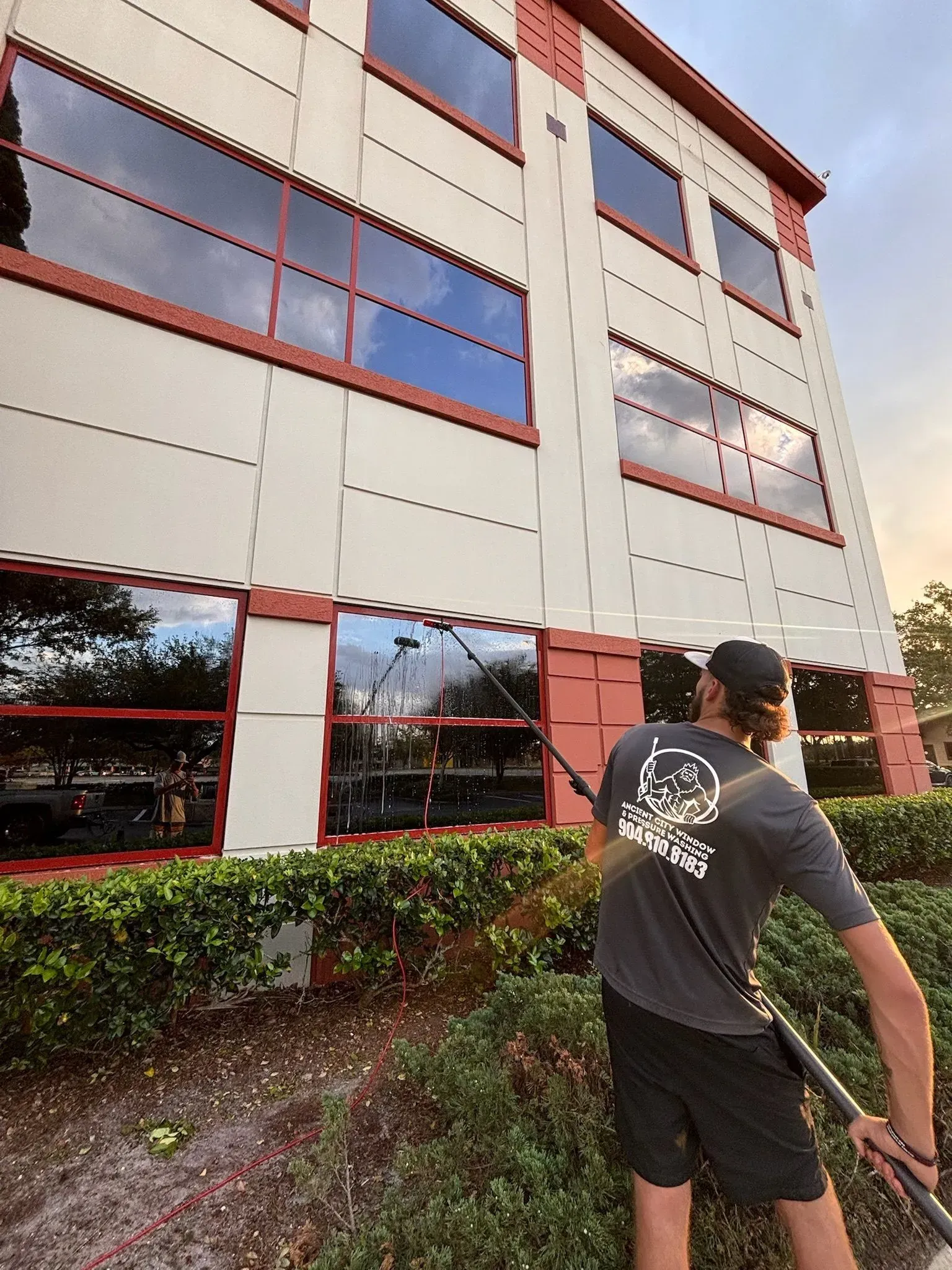 Man cleaning windows of a white office building with a long pole. Red trim, blue sky, and greenery.