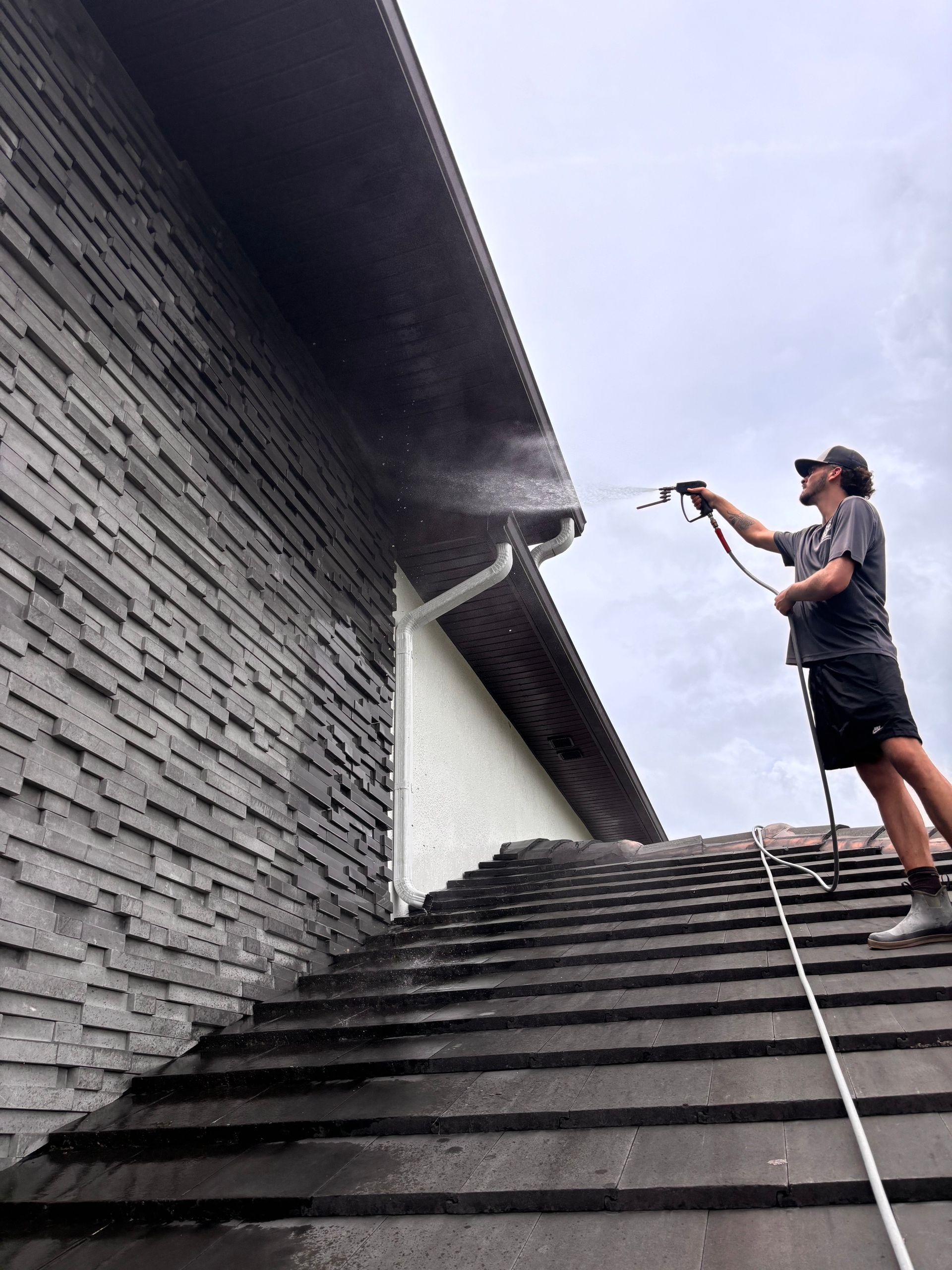 Person pressure washing a gutter on a roof. Water sprays. Gray house and roof, cloudy sky.
