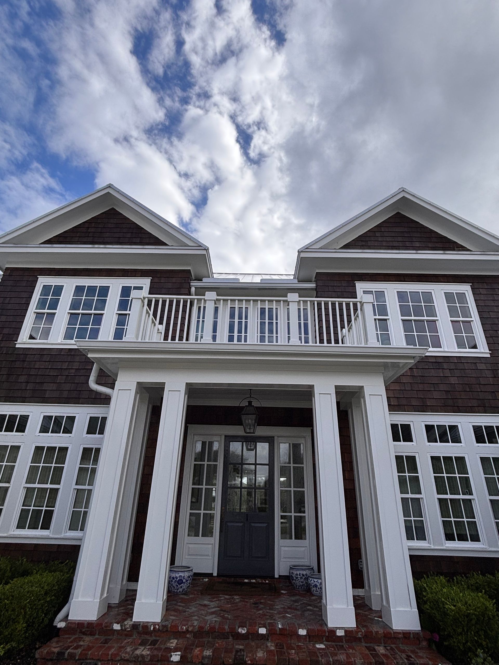Brown and white two-story house with a balcony and columns against a partly cloudy sky.