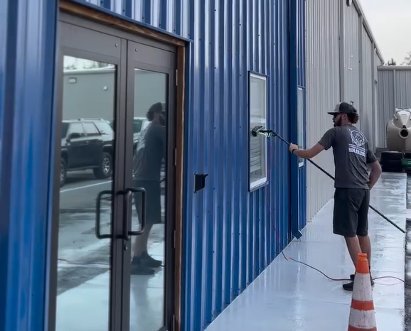 Man power washing a blue building's window; a door and a car are visible.