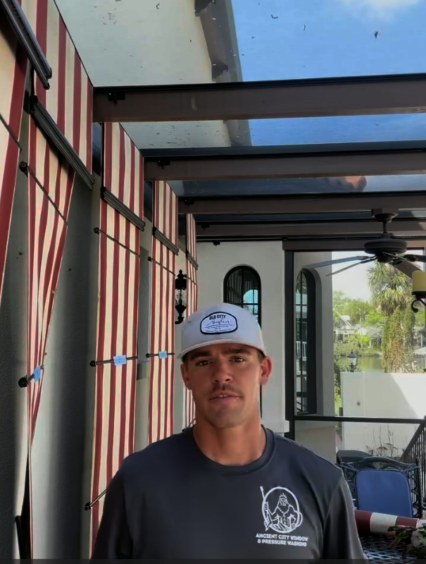 Man wearing a cap and grey shirt stands outdoors. Red and white striped curtains hang nearby.
