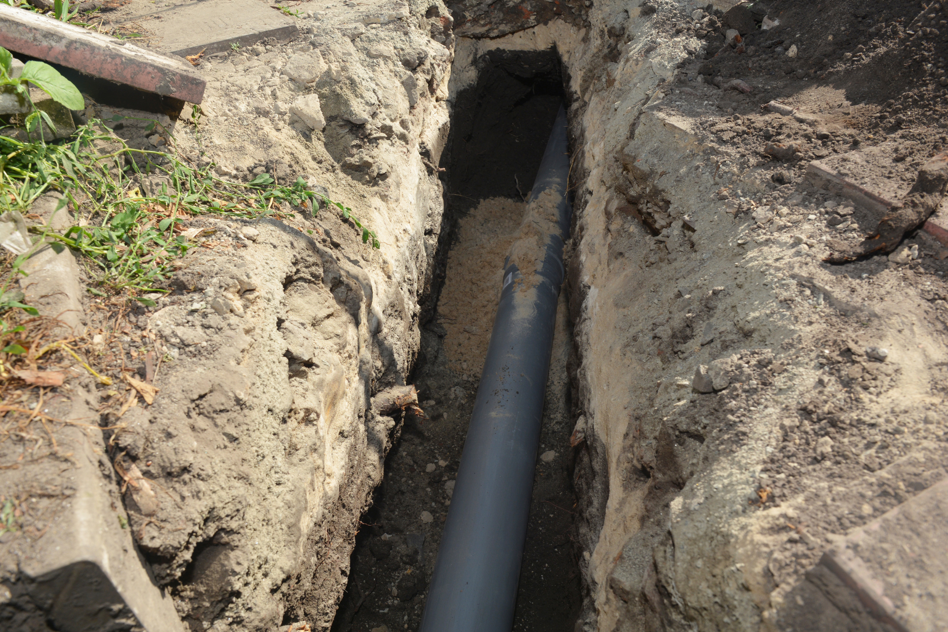 A gray pipe lies in a narrow trench dug in the ground, soil on either side.