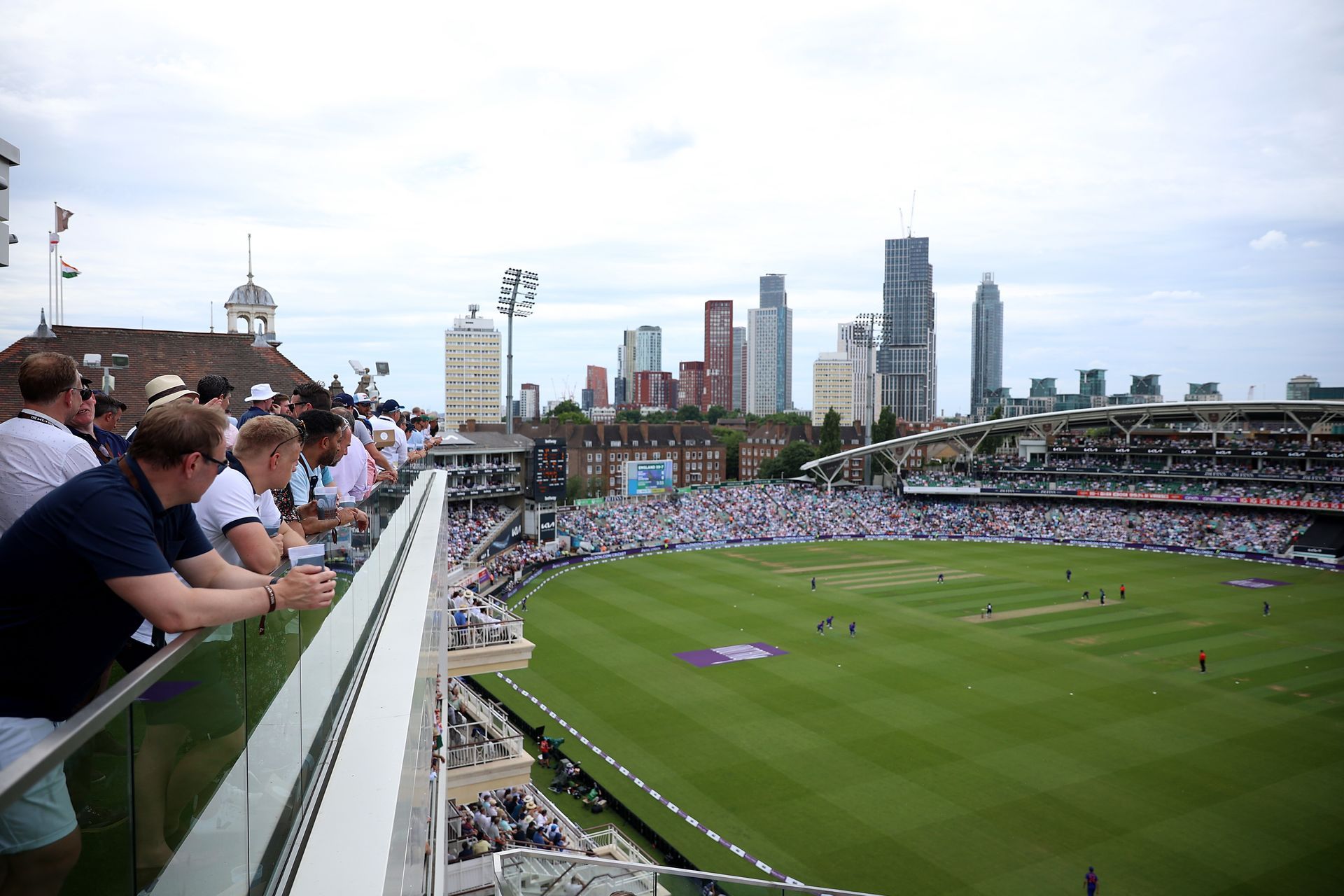 A group of people are watching a cricket match from a balcony.