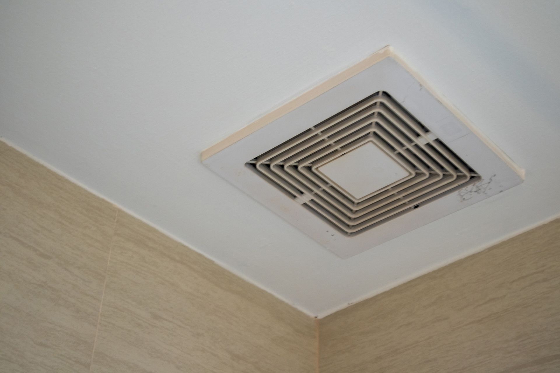 Square bathroom exhaust fan on a white ceiling, near a light beige tiled wall.