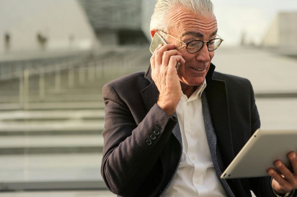 Smiling man in a dark blazer using a tablet while talking on a phone outdoors