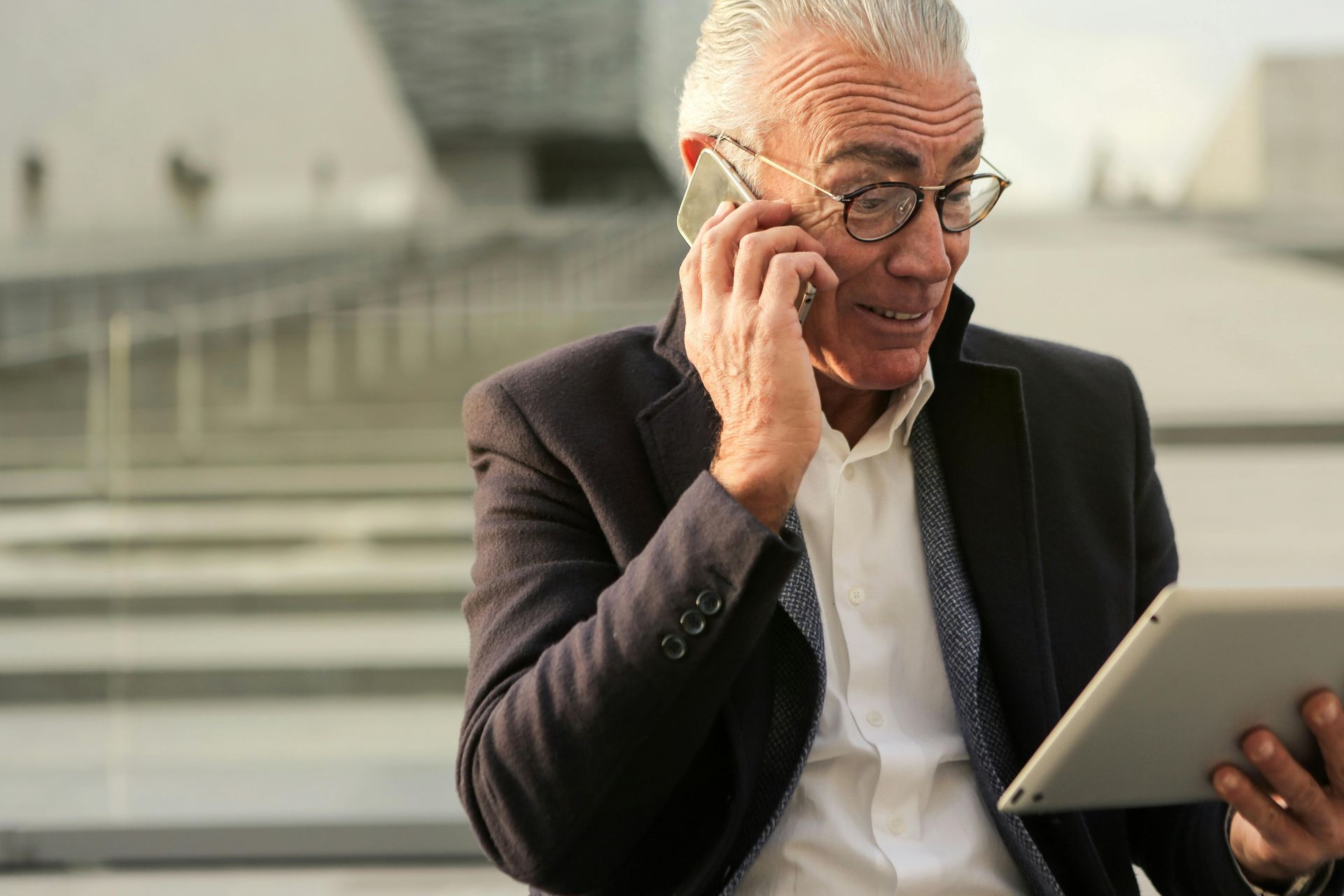 A professional in a suit and white shirt holds a tablet while talking on a smartphone in a modern, outdoor plaza.