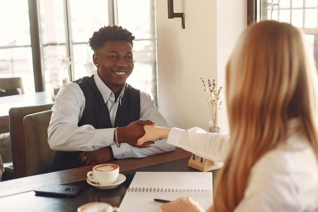Two people shaking hands across a café table, with coffee and a notebook in front of them.