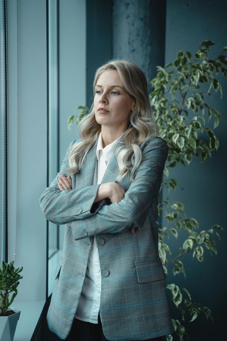 Woman in a blue patterned blazer standing with arms crossed by a window and leafy plant