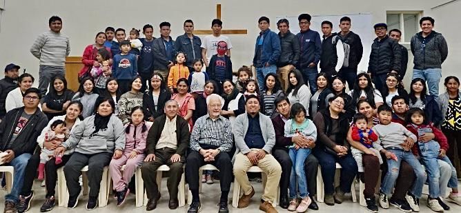 Group photo of a large gathering of people in a room, some smiling, with a cross in the background.