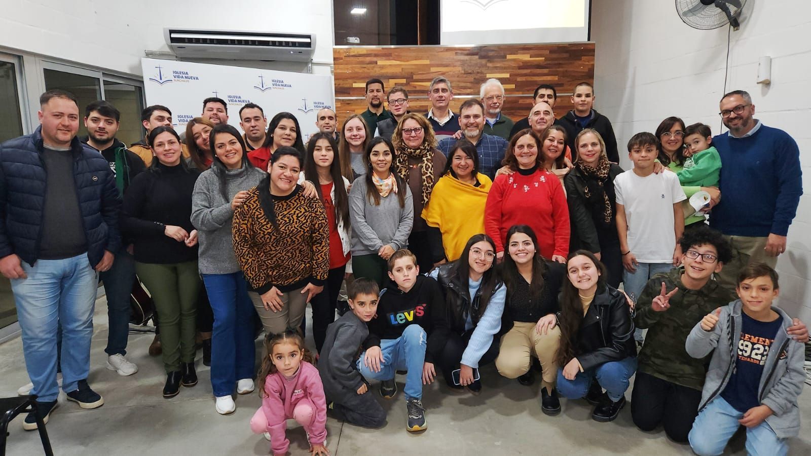 Group photo: diverse people smiling indoors. Some stand, some kneel. Wooden backdrop.