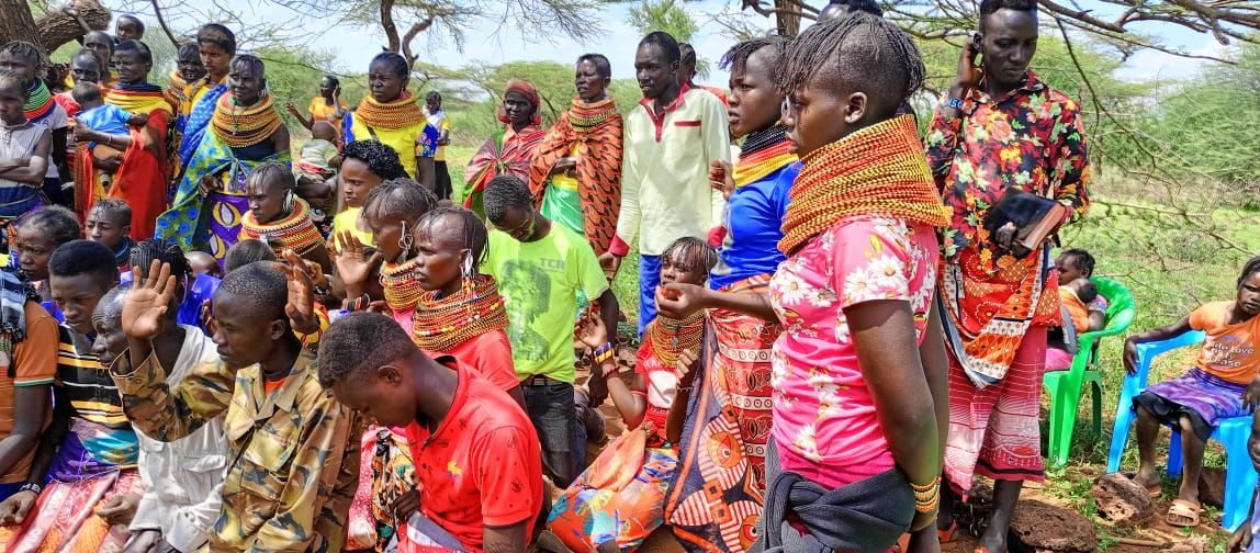 Group of people, some kneeling, in colorful attire, outdoors under trees.