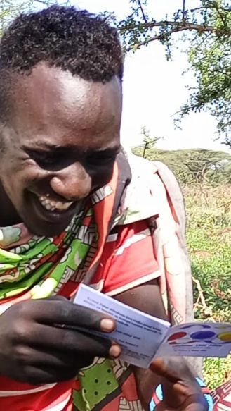 Man in traditional attire smiles while looking at a small pamphlet outdoors, with a green and brown background.