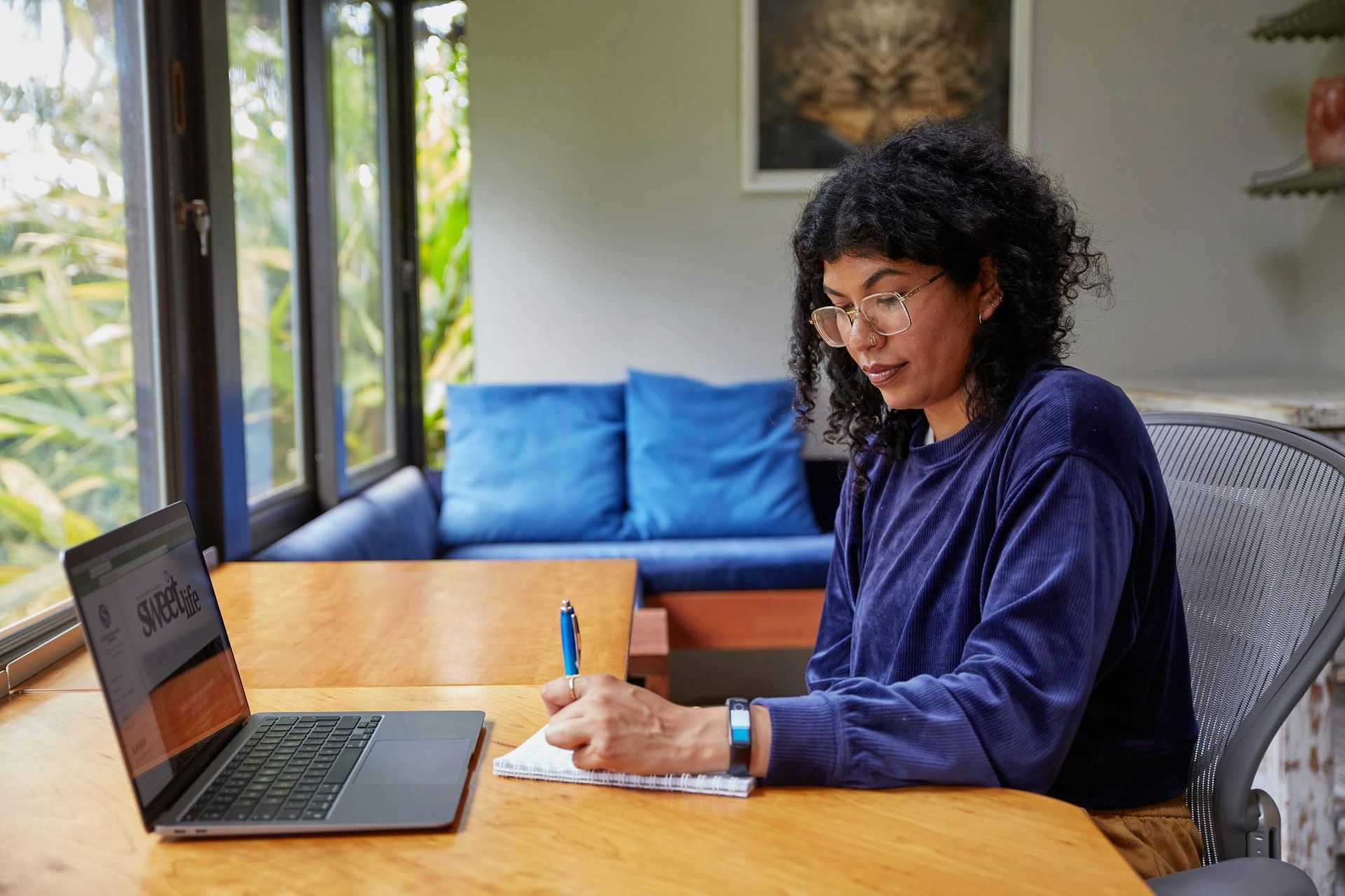 A woman in glasses writing in a notebook at a table with a laptop near a window.