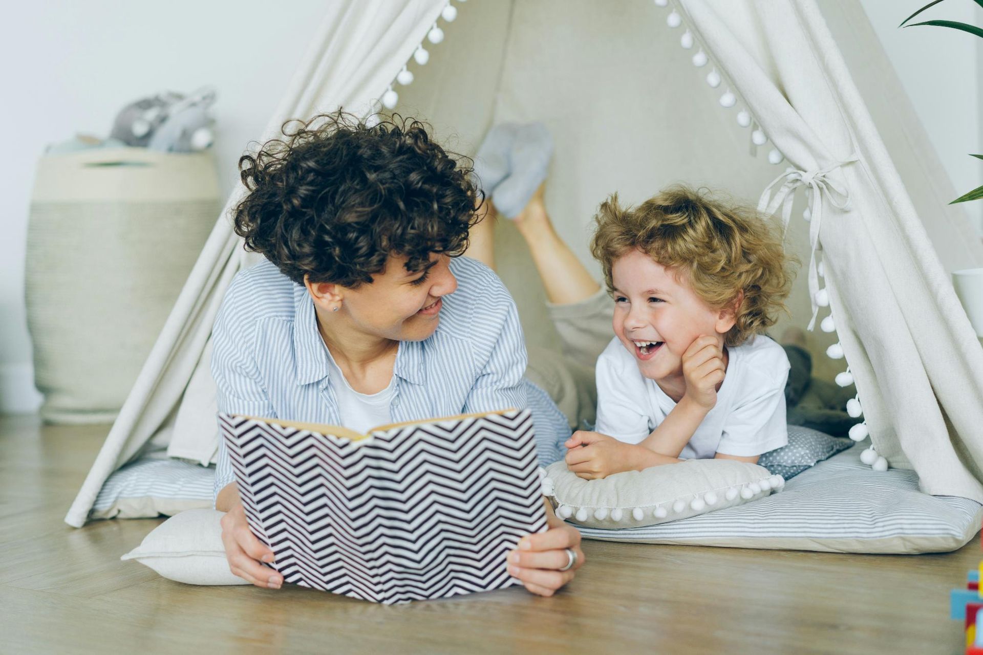 An adult and a child are lying inside a cozy indoor tent as they read a book.