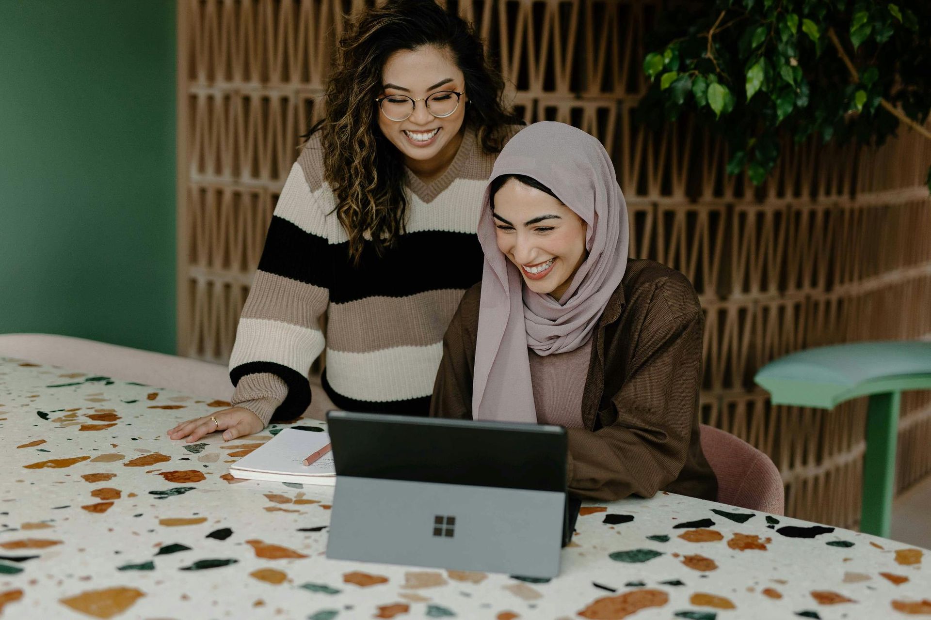 Two women smiling and collaborating while working on a laptop in a modern, stylish workspace.