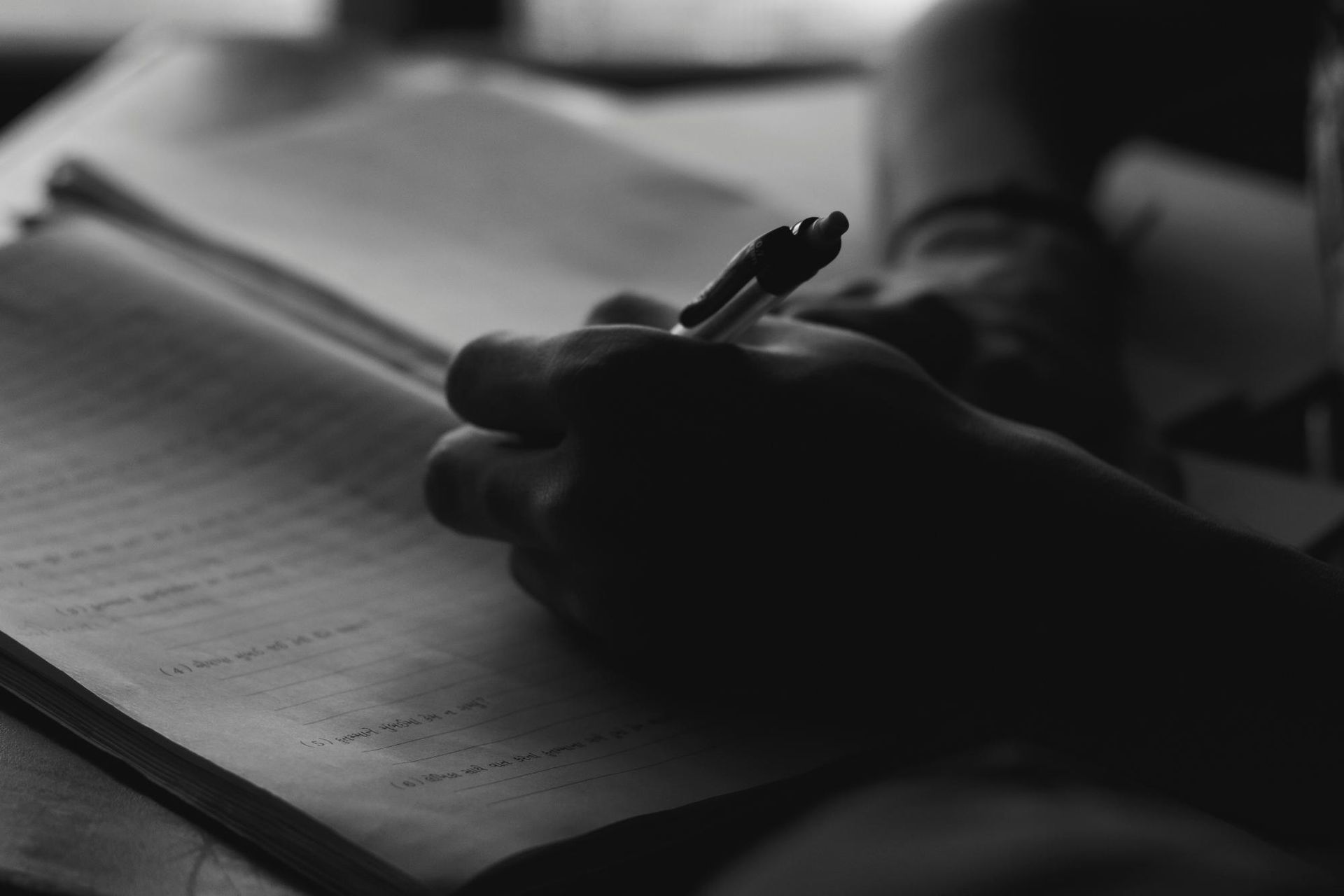 A close-up of a hand writing in a notebook, captured in moody black and white.