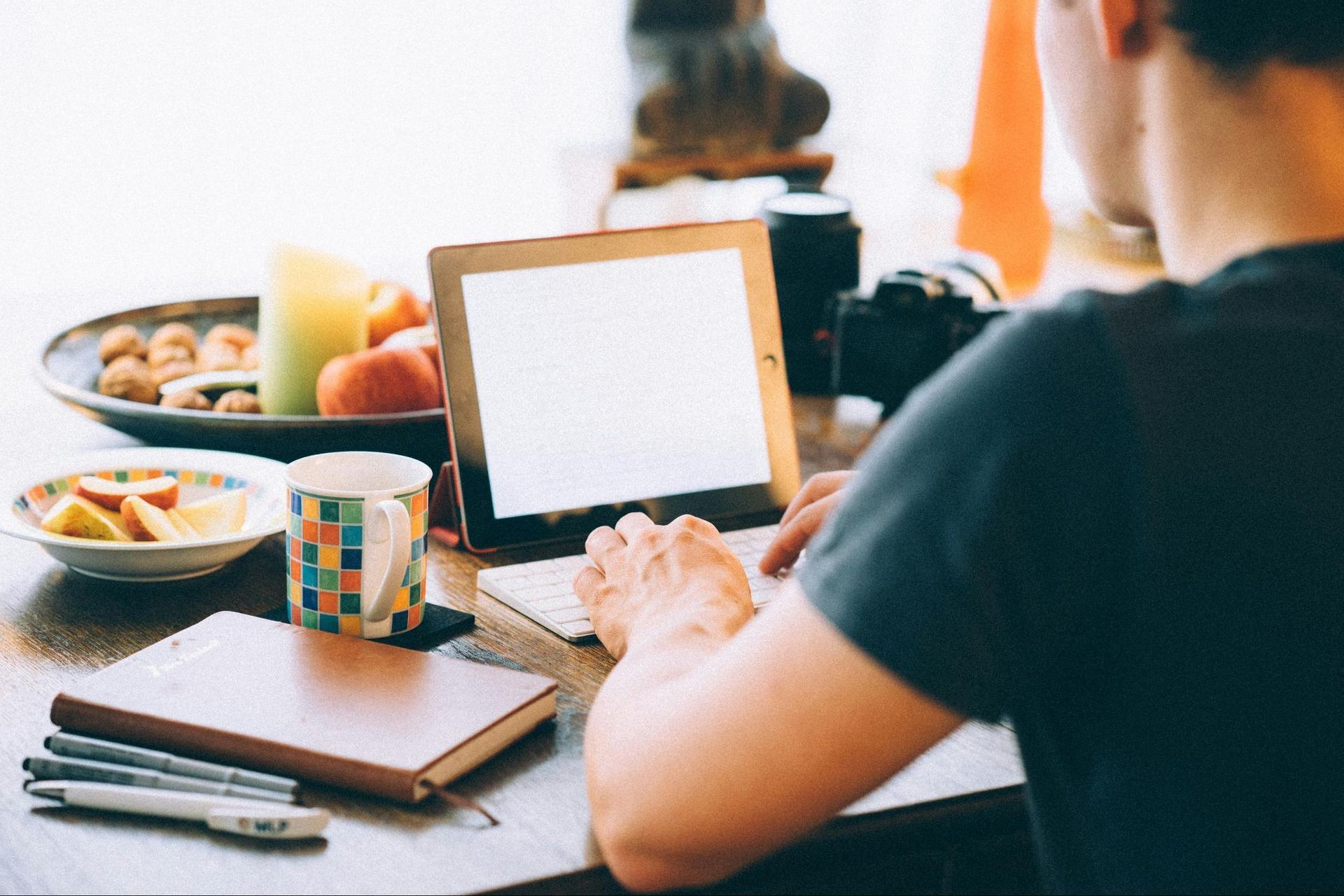 A person working on a tablet at a table, surrounded by a notebook, pens, a cup of tea, and snacks.