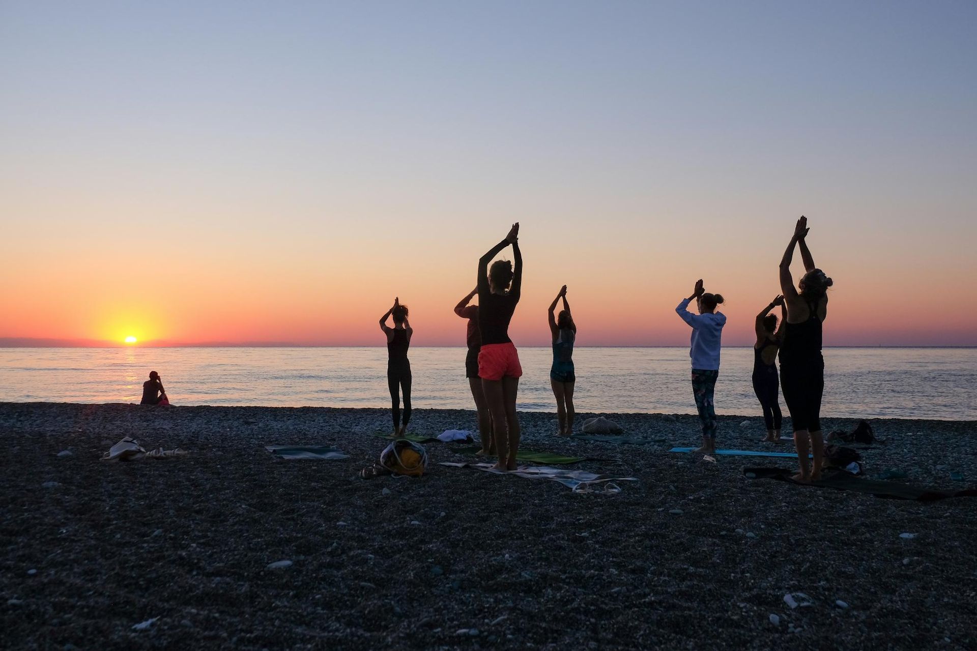 
A group of people practices yoga on a beach at sunset, with the sun low on the horizon and the calm