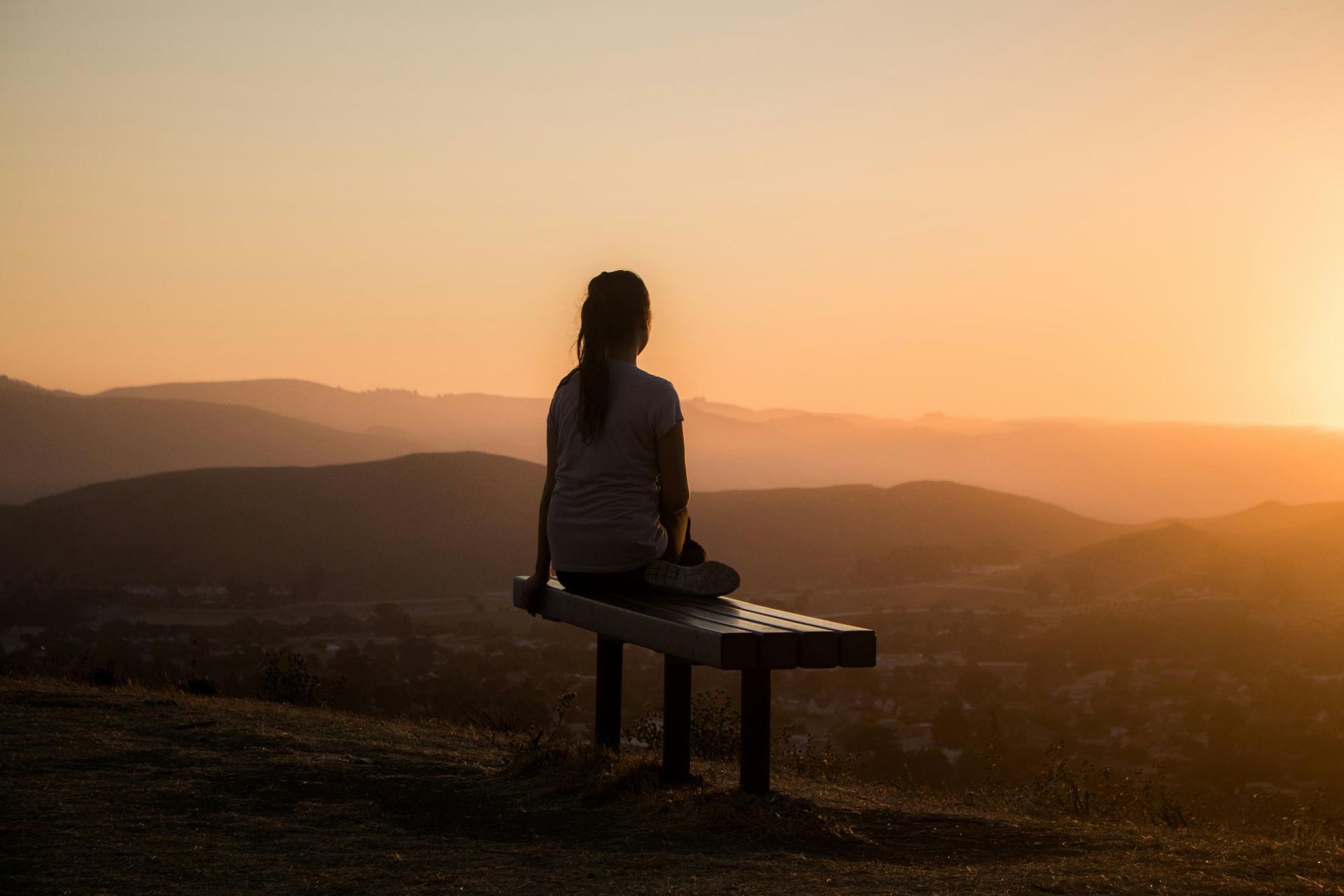 A person sits on a bench overlooking a scenic landscape of hills during a sunset