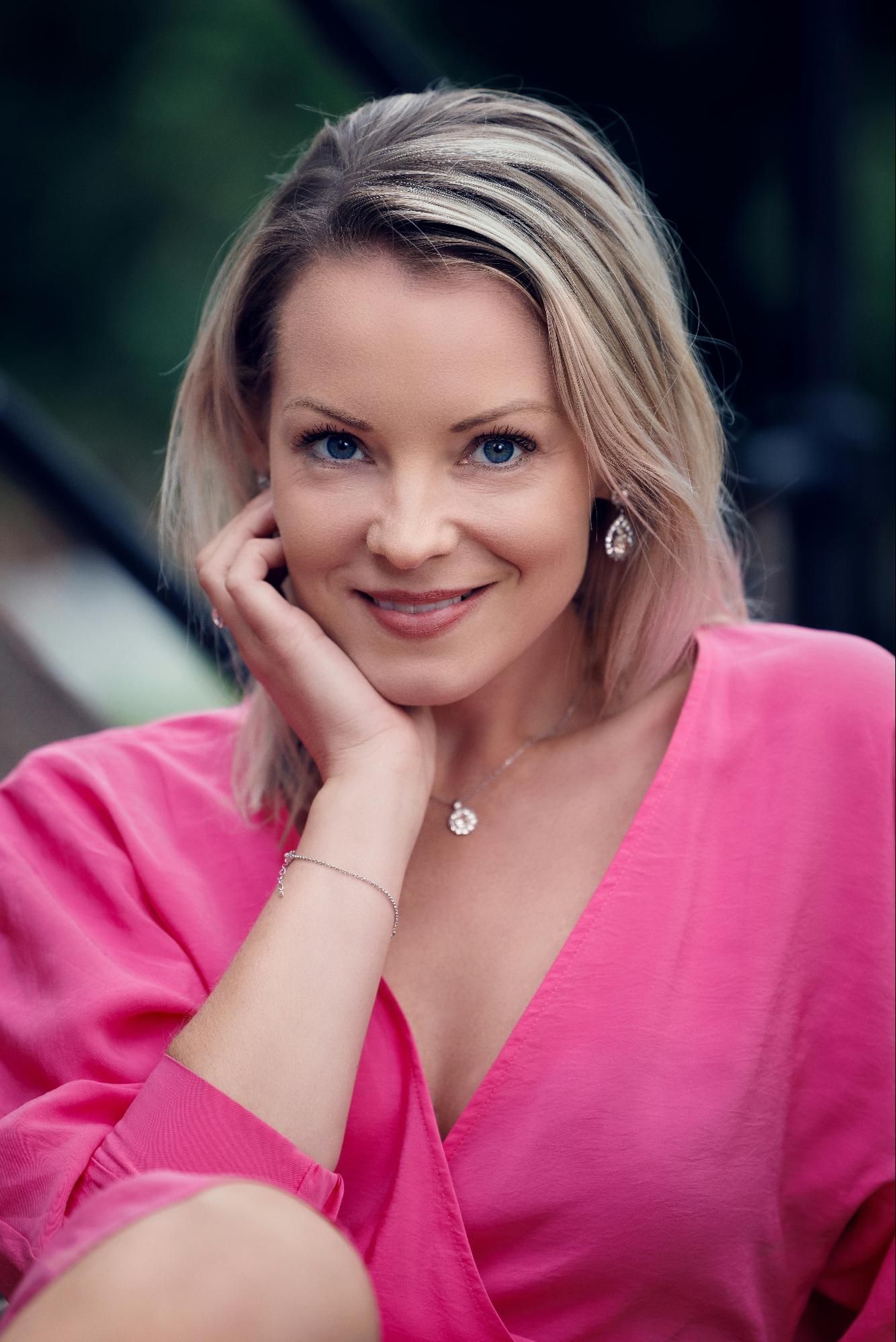 A woman with blonde hair smiles softly, resting her hand on her chin wearing bright pink shirt