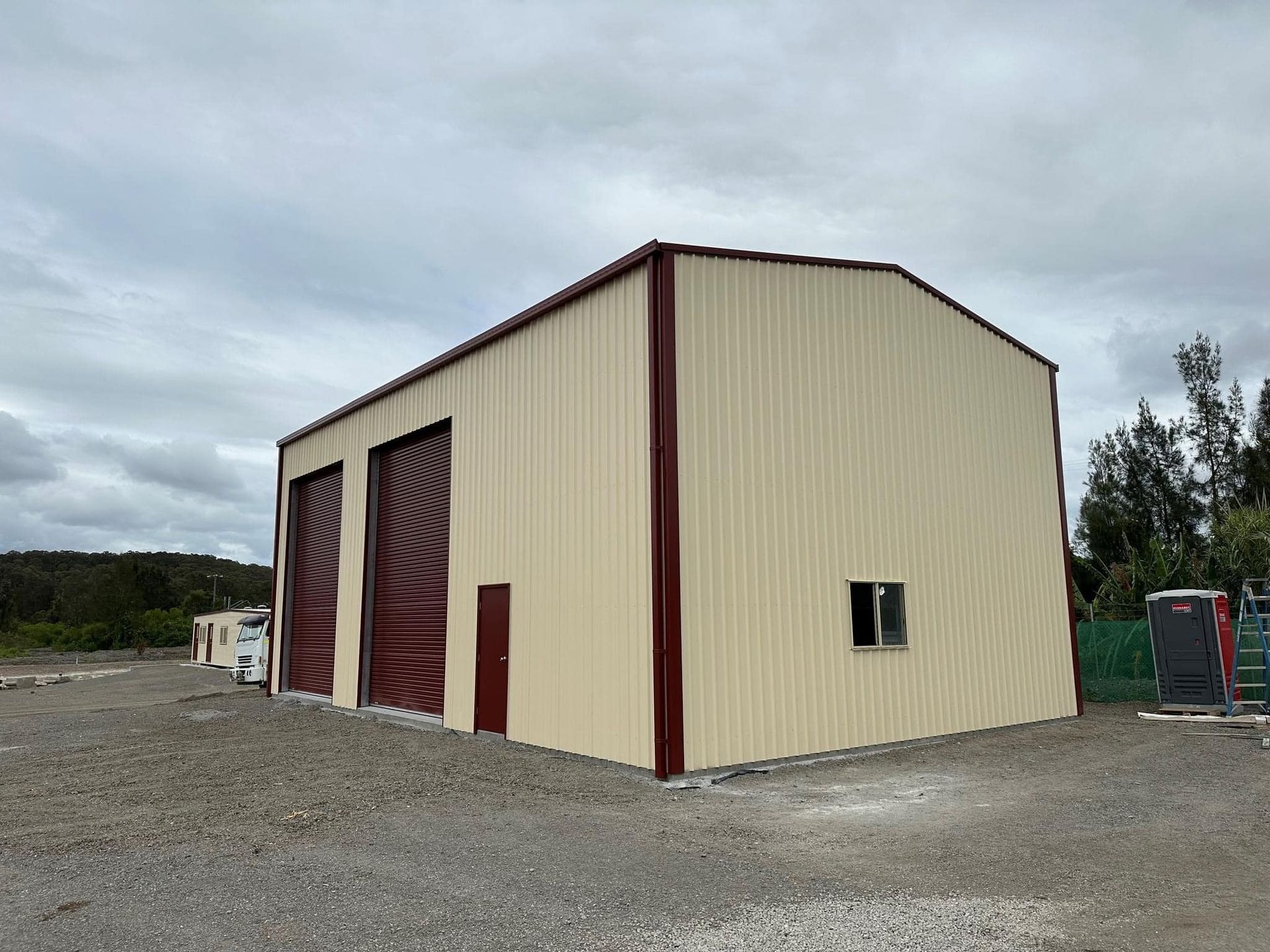 A large metal building with two red garage doors is sitting on top of a gravel lot.