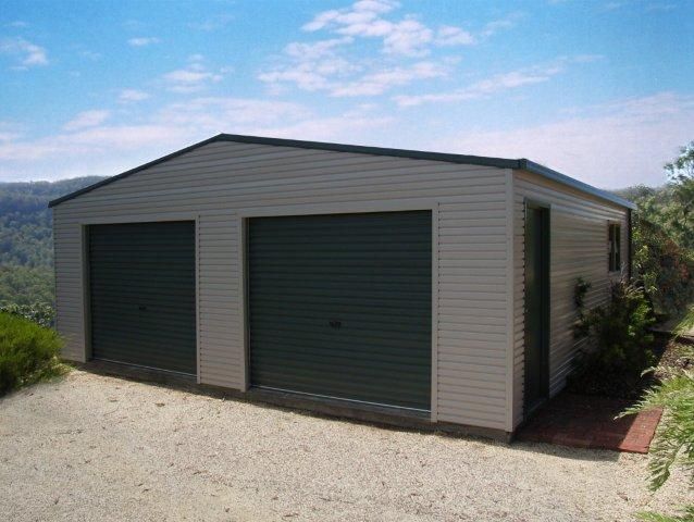 A white garage with two green garage doors is sitting on top of a gravel driveway.