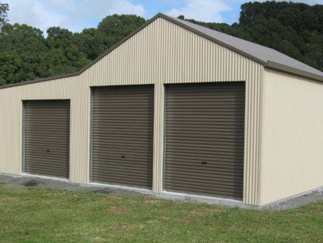 A garage with three garage doors is sitting in the middle of a grassy field.