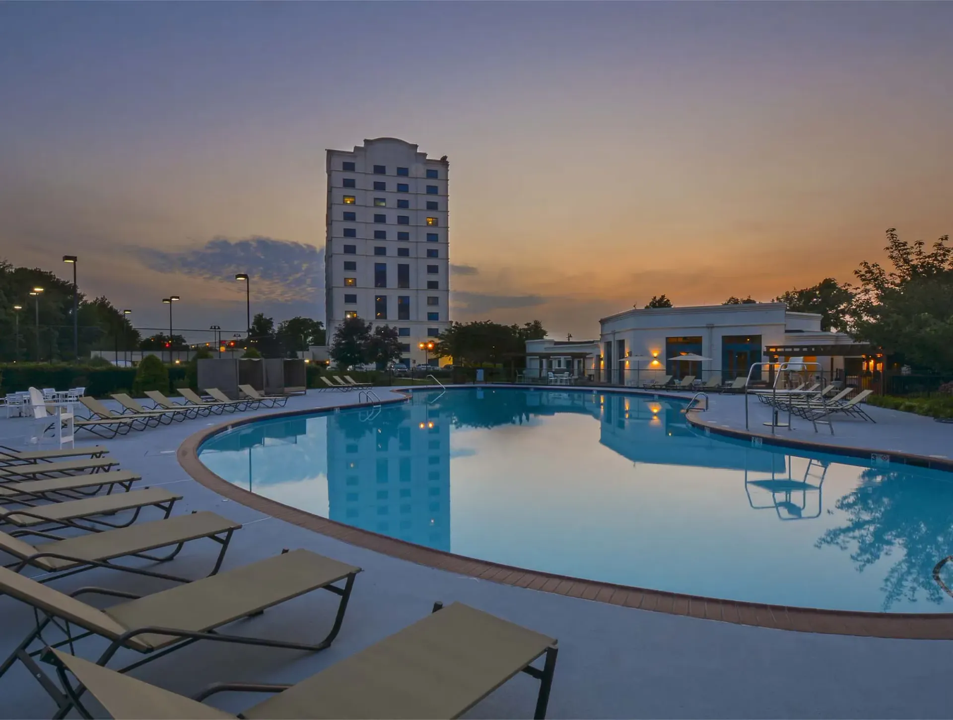 Outdoor pool with lounge chairs surrounding it at dusk, with a building in the background.