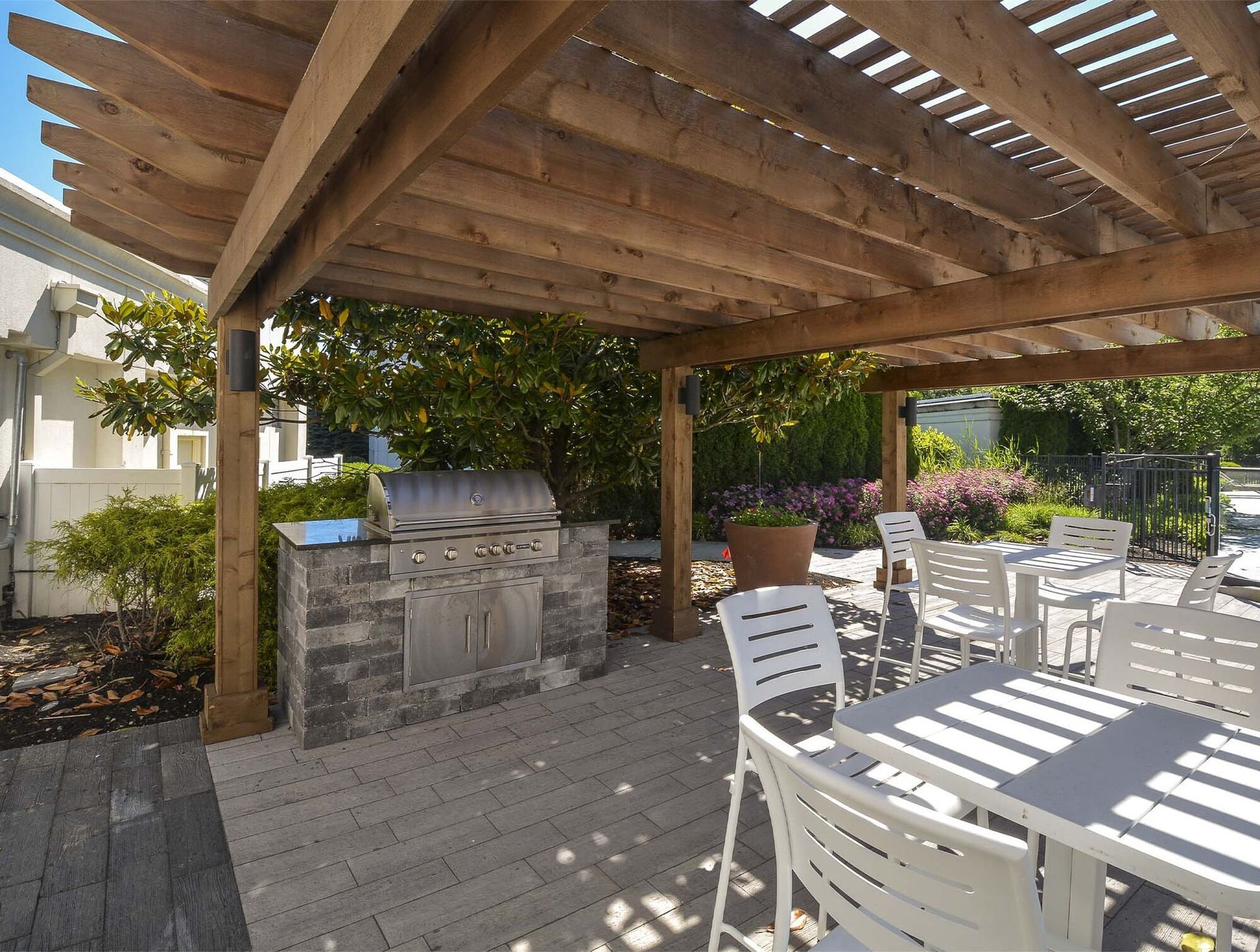 Outdoor community patio under a wooden pergola with a built-in grill and white dining tables.