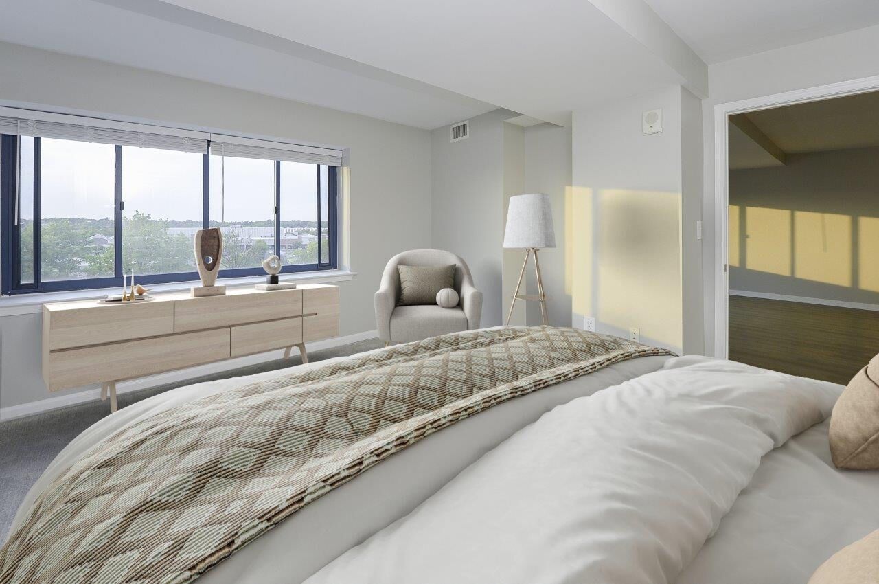 Modern bedroom in a light-filled apartment with a low wood dresser, chair, and large window.