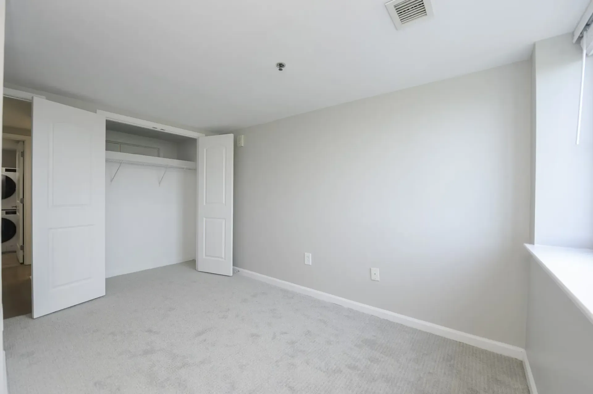 Empty bedroom with a white closet and visible laundry appliances in the hallway.