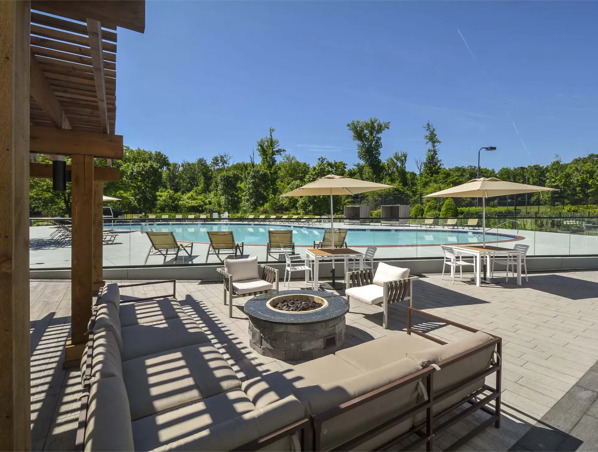 Outdoor pool deck with lounge seating, tables, umbrellas, and a glass barrier.