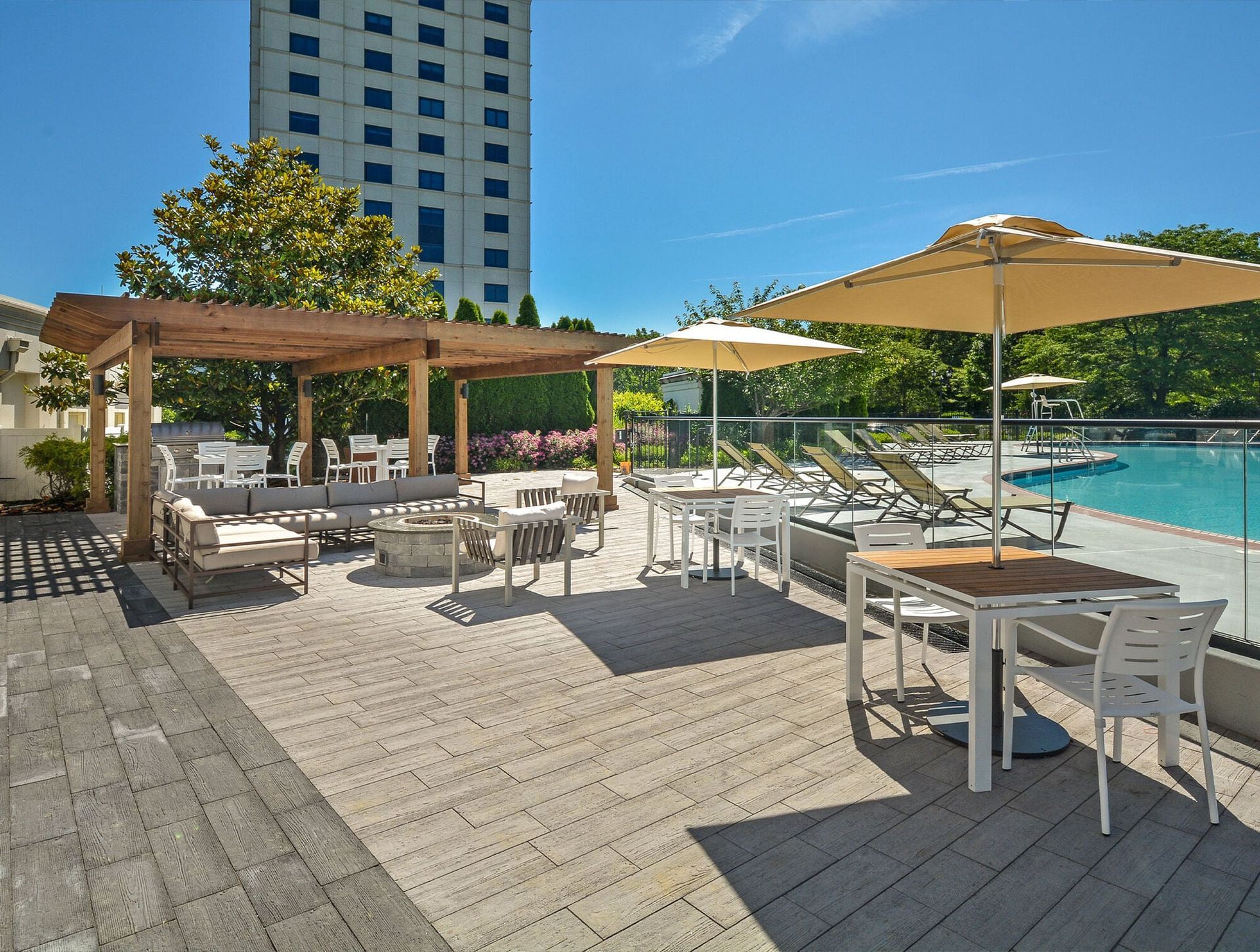 Outdoor pool deck with lounge chairs, tables, and umbrellas beside a glass railing.