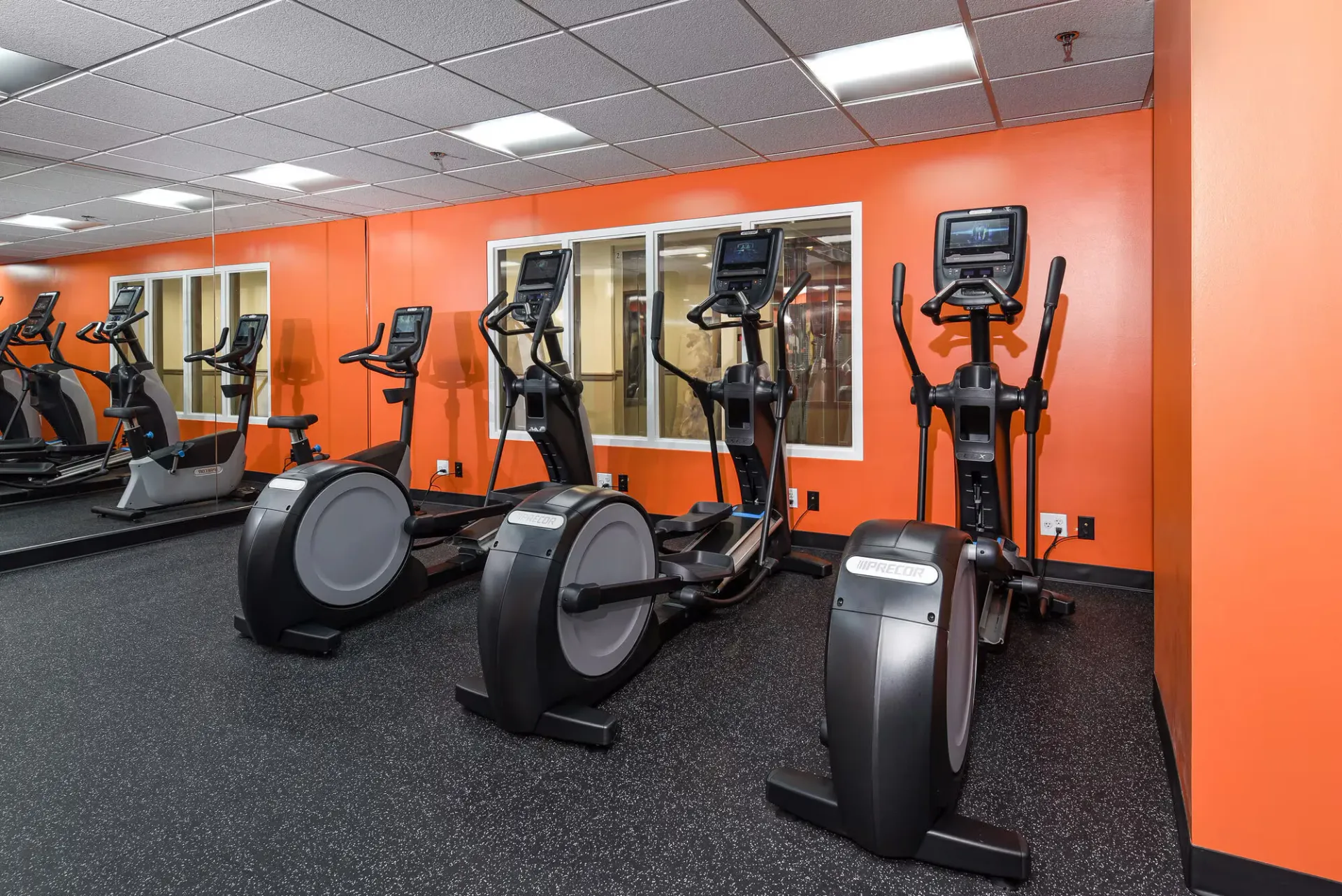 Cardio machines lined up in an orange-walled gym.