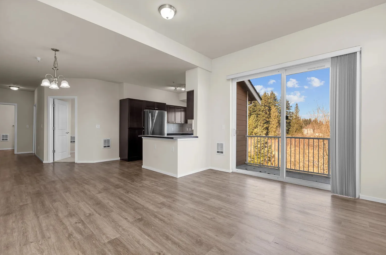 Open-concept apartment with wood floors, kitchen, and sliding glass door to a balcony overlooking trees.
