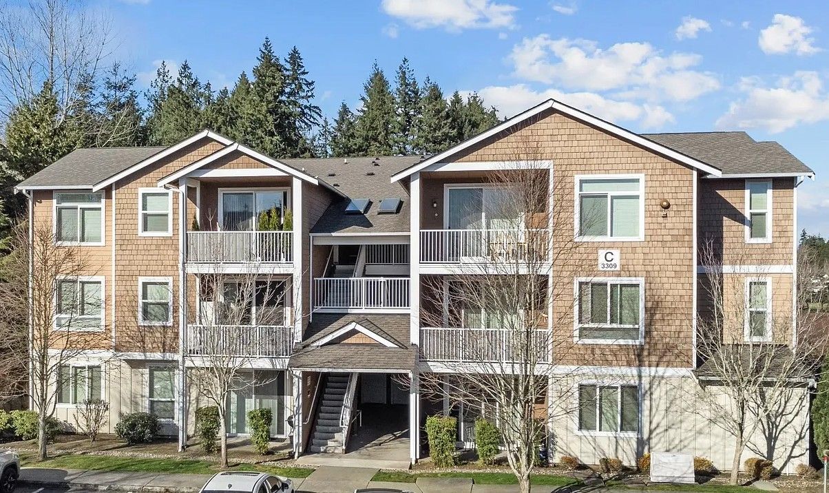 Three-story apartment building with brown siding, white balconies, and a dark roof, set against a backdrop of trees.