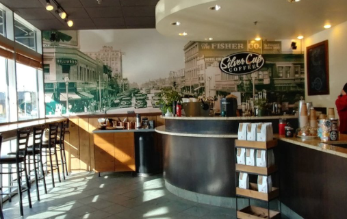 Interior of a coffee shop: counter, tables, mural of city scene, and packaged coffee.