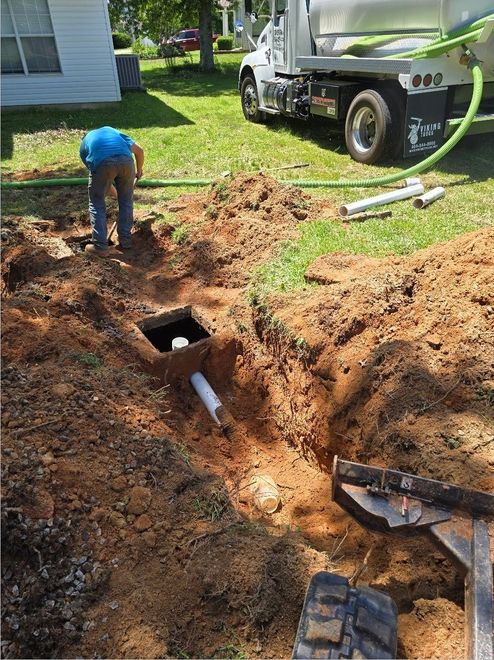 Man working on a septic system. Open dirt pit, white truck with hose, green grass, house in background.