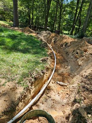 Trench dug in a yard with white pipe installed. Green grass on the left and trees in the background.