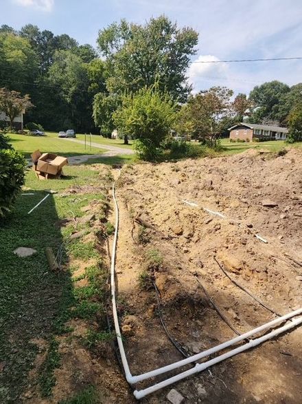 Trench with white pipes laid in it, surrounded by dirt and grass, trees in the background, sunny day.