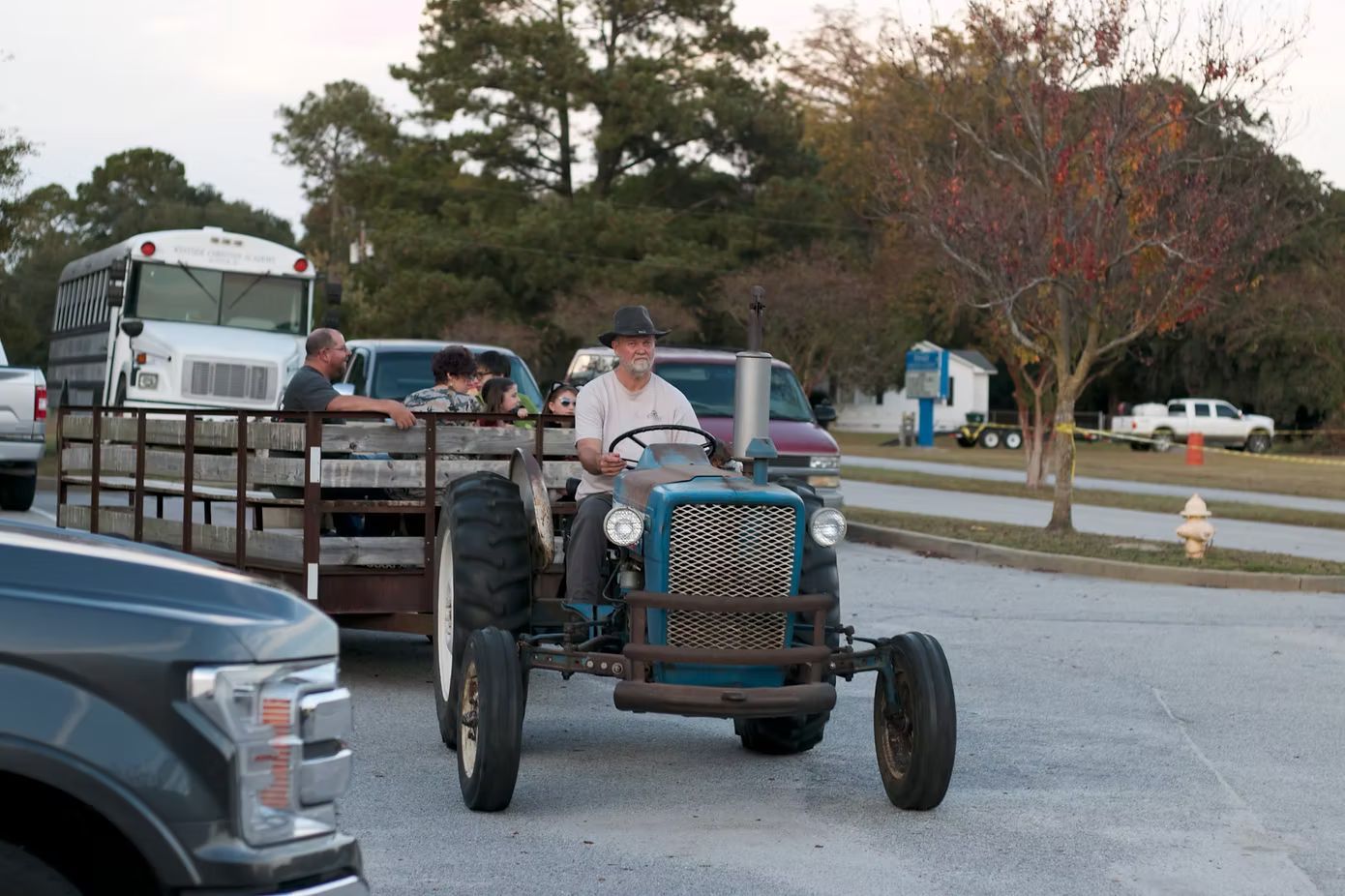 Man driving a blue tractor pulling a wagon with people; cars and bus parked nearby.
