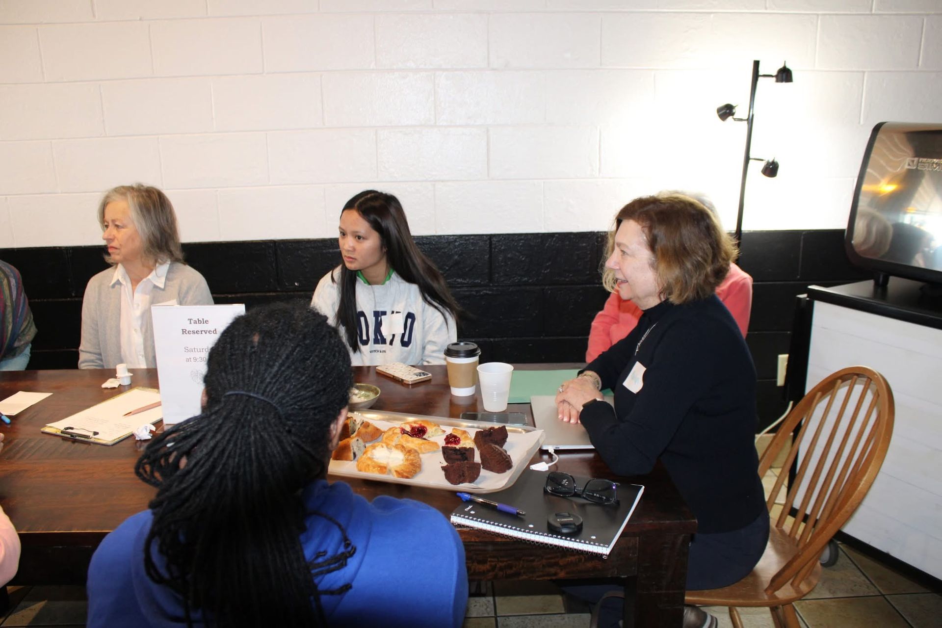 People seated at a table, discussing. Snacks and documents present. Brightly lit room with a black and white wall.