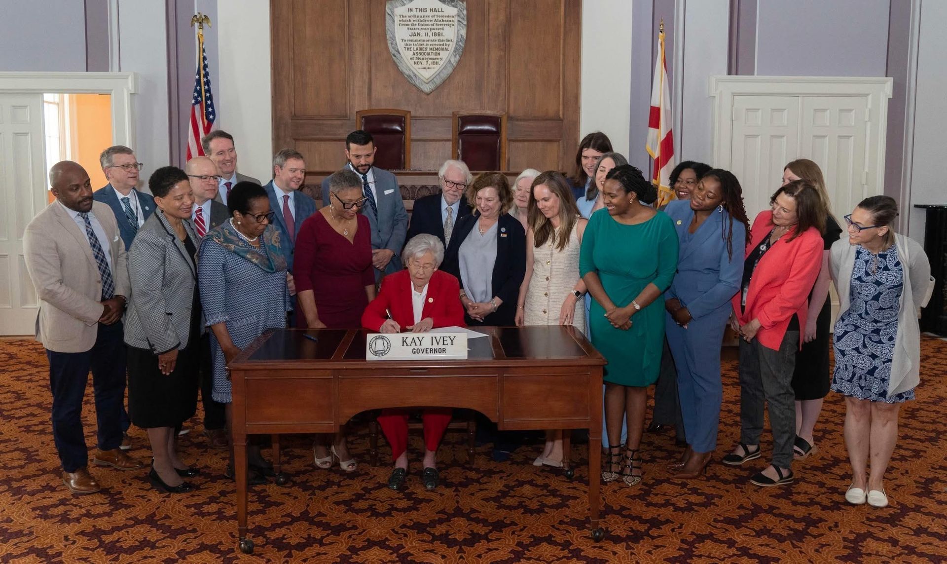 Woman signing document at a wooden desk, surrounded by a group of people in a room with a state seal.