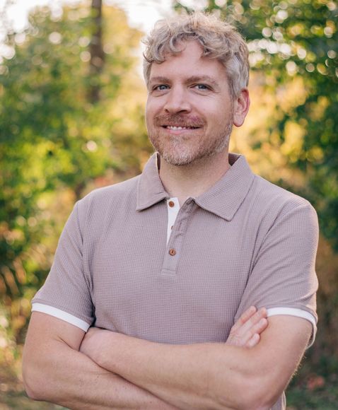 A man with curly hair and a beard is standing with his arms crossed and smiling.