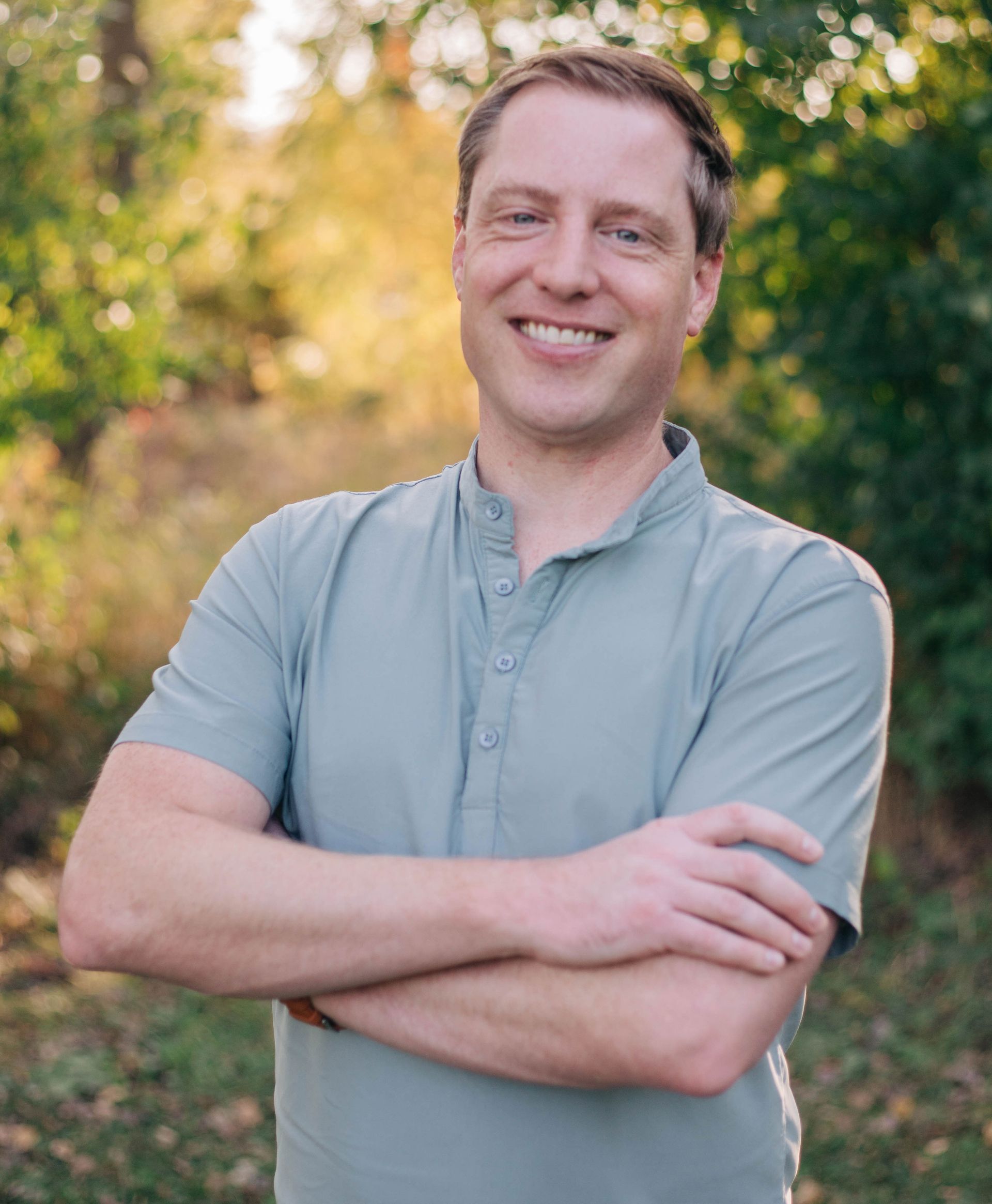 A man in a grey shirt is standing with his arms crossed and smiling.