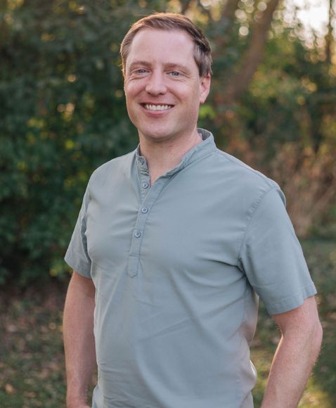 A man in a green shirt is standing in the woods with his hands in his pockets.